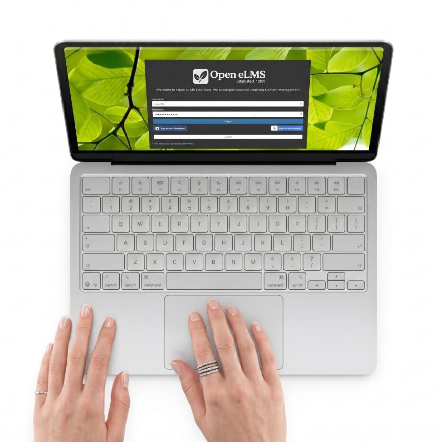 A top-down, minimalist shot of a silver laptop centered against a completely seamless, solid white background. The laptop's screen displays a login page titled 'OPEN eLMS' against a vibrant background of bright green leaves. A pair of light-skinned hands rests on the lower portion of the laptop; the left hand is placed near the edge, wearing a simple band on the ring finger, while the right hand rests on the trackpad, wearing a thick, textured silver ring.